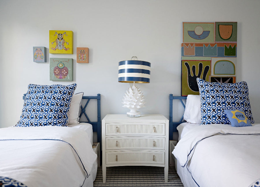 Two beds with blue and white patterned pillows, a nightstand with a lamp, and colorful artwork on the wall.